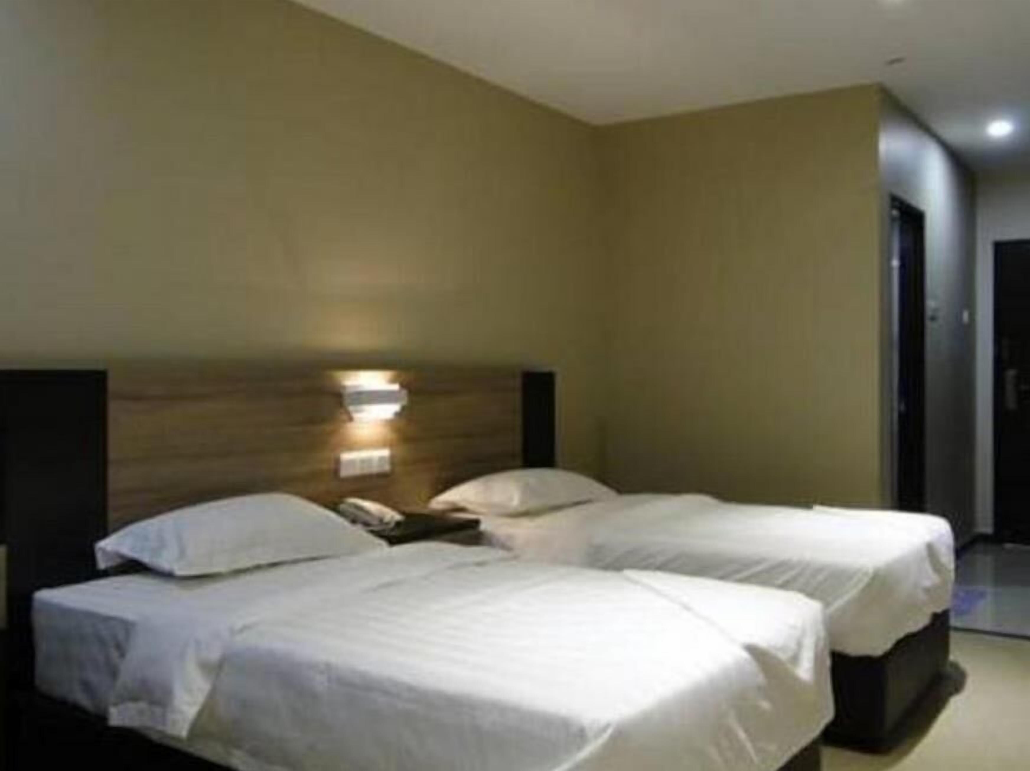A hotel room with two single beds, a wooden headboard, a bedside table with a lamp, and a door leading to a hallway.