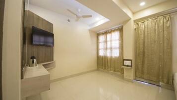 A spacious room with a flat-screen TV mounted on a wooden panel, a ceiling fan, a large window with curtains, and a tiled floor.