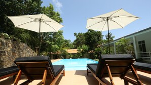 A poolside area with wooden lounge chairs and two umbrellas, surrounded by greenery and a stone wall. - Beach Grove Villas (Unawatuna)