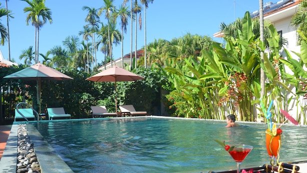 A pool area with lounge chairs, umbrellas, and tropical plants. - Hoi An Red Frangipani Villa (Da Nang)
