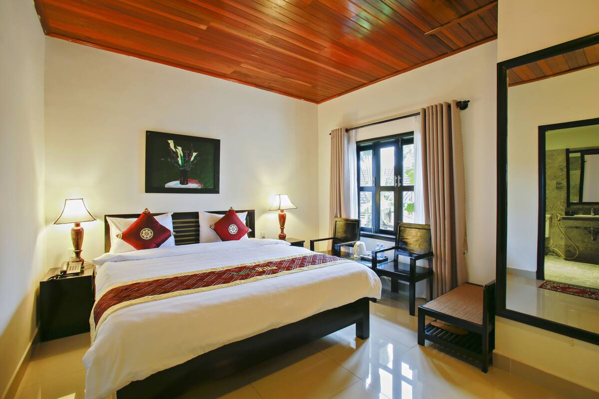 A hotel room with a large bed, two bedside lamps, a television, a small table with a chair, and a window with curtains.