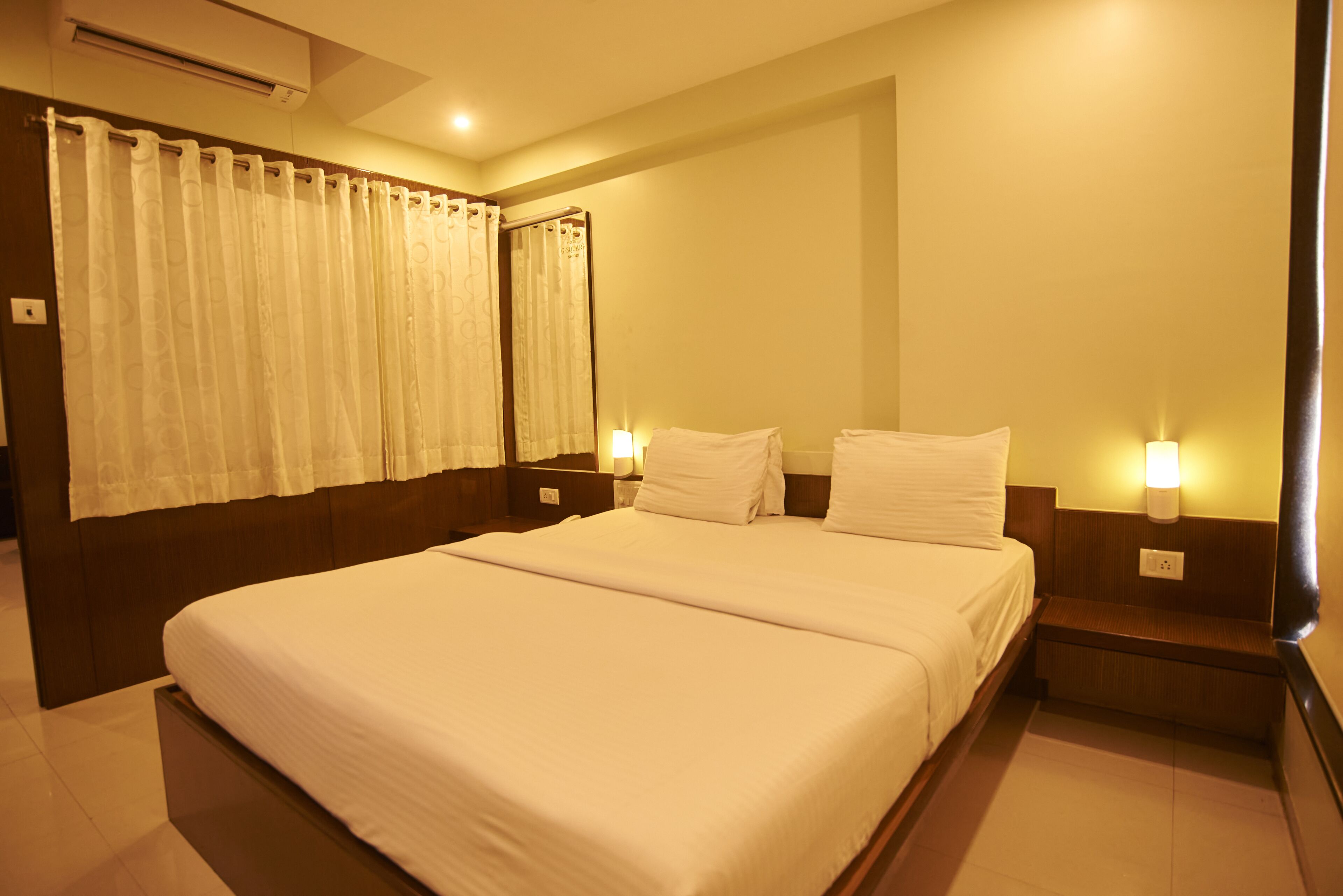 Een hotelkamer met een groot bed, twee nachtkastjes met lampen, een raam met gordijnen en een airconditioning.