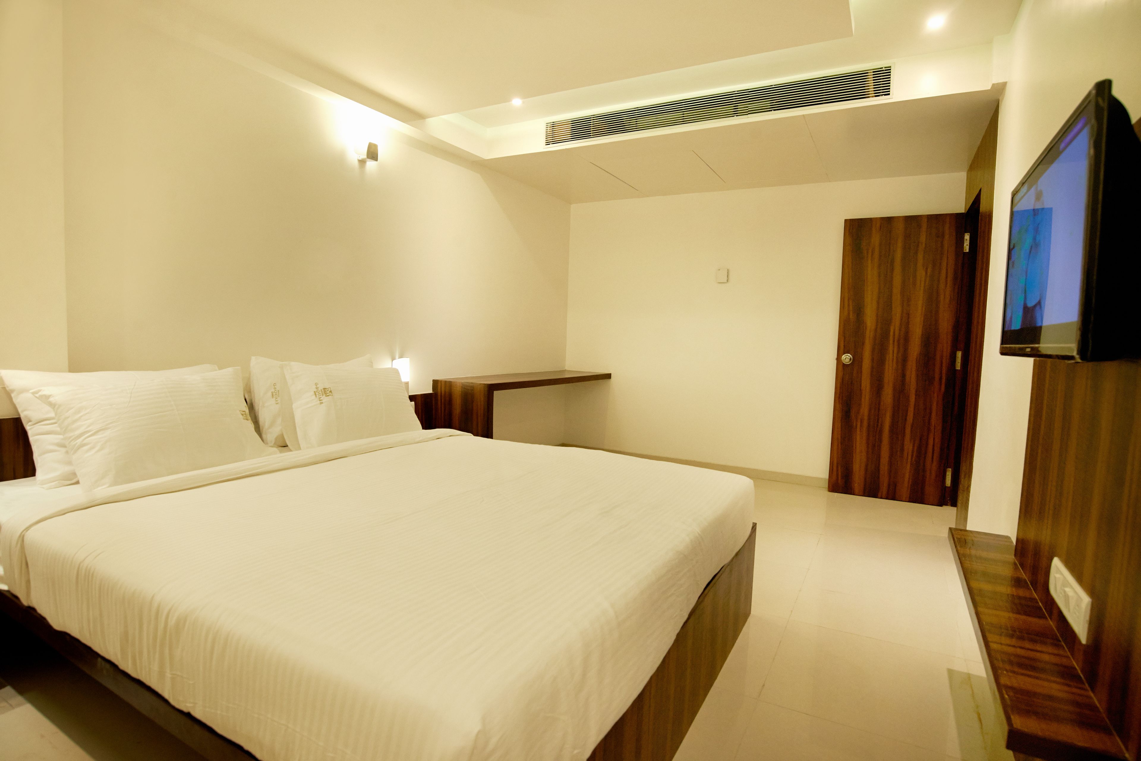 A modern bedroom with a large bed, a wooden nightstand, a TV mounted on the wall, and a door leading to another room.