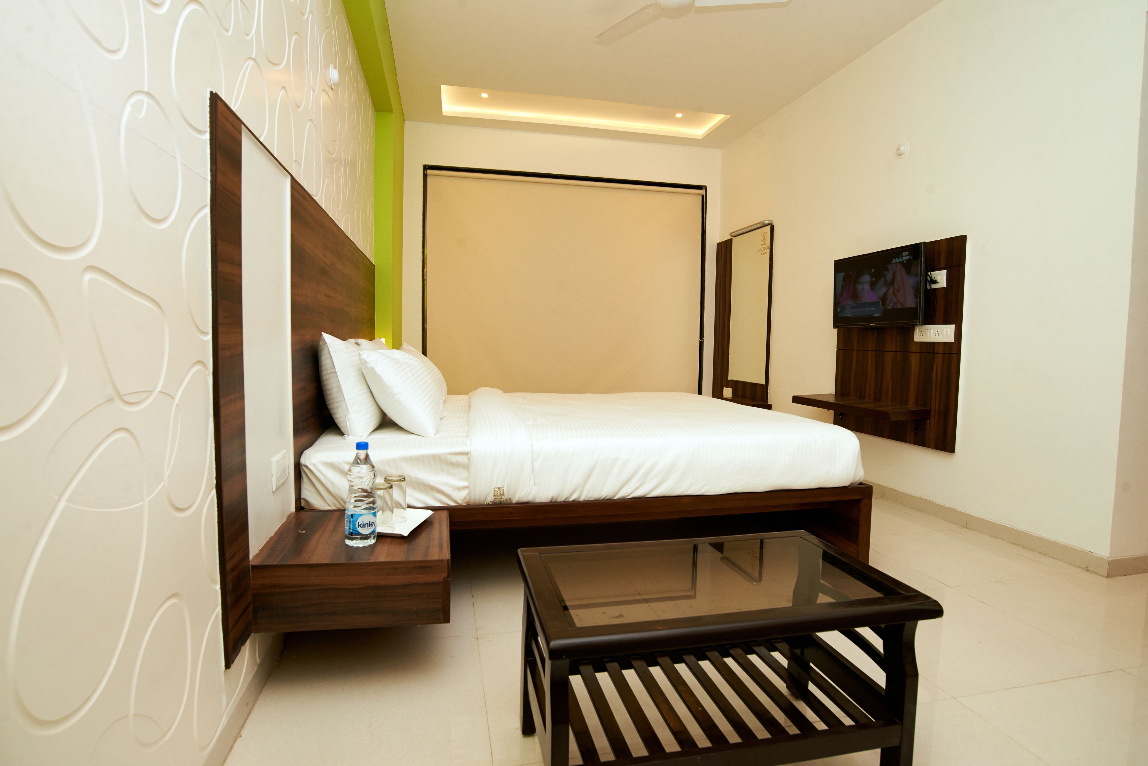 Una habitación de hotel moderna con una cama, una mesita de cristal, una mesita de noche con una botella y un televisor en la pared.