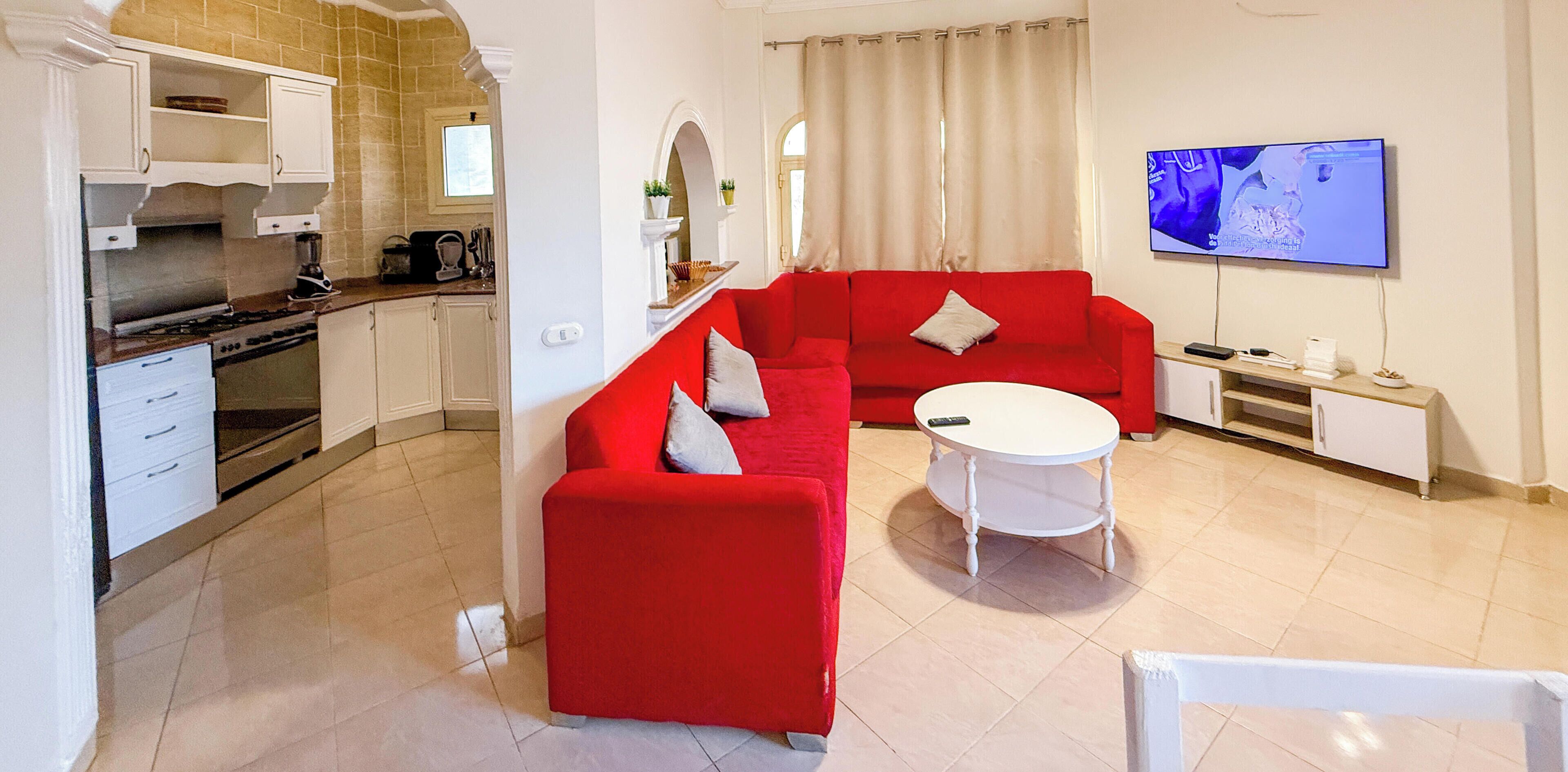 Un espace de vie compact comprenant un coin cuisine, un canapé rouge, une table basse blanche et un téléviseur à écran plat fixé au mur.