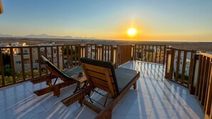 A wooden deck with two lounge chairs and a small table, overlooking a cityscape and mountains during sunset. - Green Garden Resort (Hurghada)