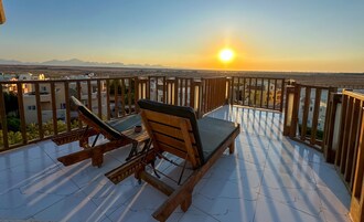 A wooden deck with two lounge chairs and a small table, overlooking a cityscape and mountains during sunset.