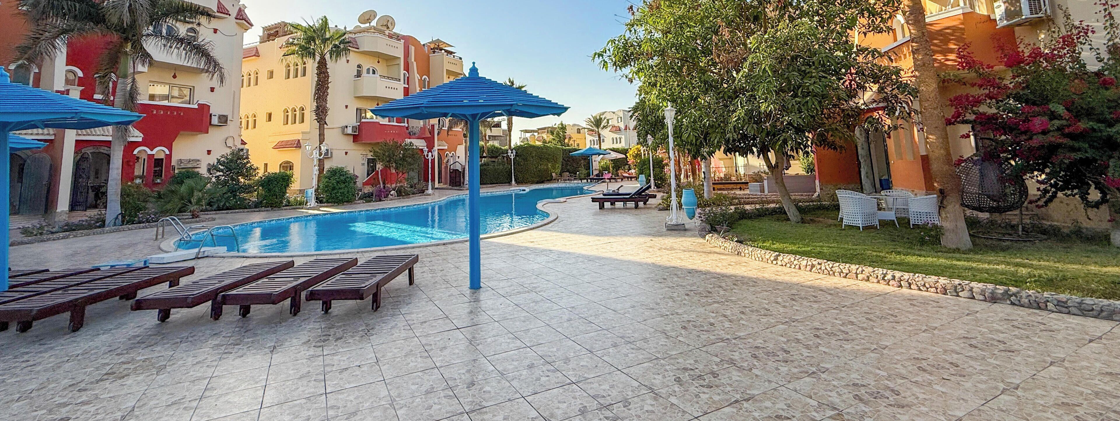 A swimming pool area with lounge chairs and umbrellas, surrounded by buildings with balconies and satellite dishes.
