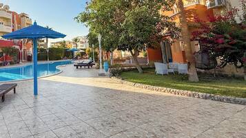 A swimming pool area with lounge chairs and umbrellas, surrounded by buildings with balconies and satellite dishes.