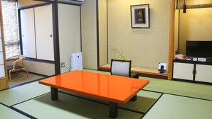 Una habitación japonesa tradicional con suelo de tatami, una mesa baja anaranjada, un televisor y un refrigerador pequeño.