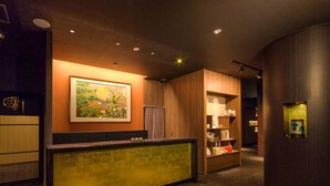Zona de recepción de un hotel con mostrador, un cuadro en la pared y una estantería de madera con varios objetos.