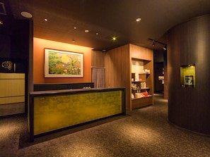 A hotel reception area with a reception desk, a painting on the wall, and a wooden bookshelf with various items. - Kanazawa Chaya (Kanazawa)