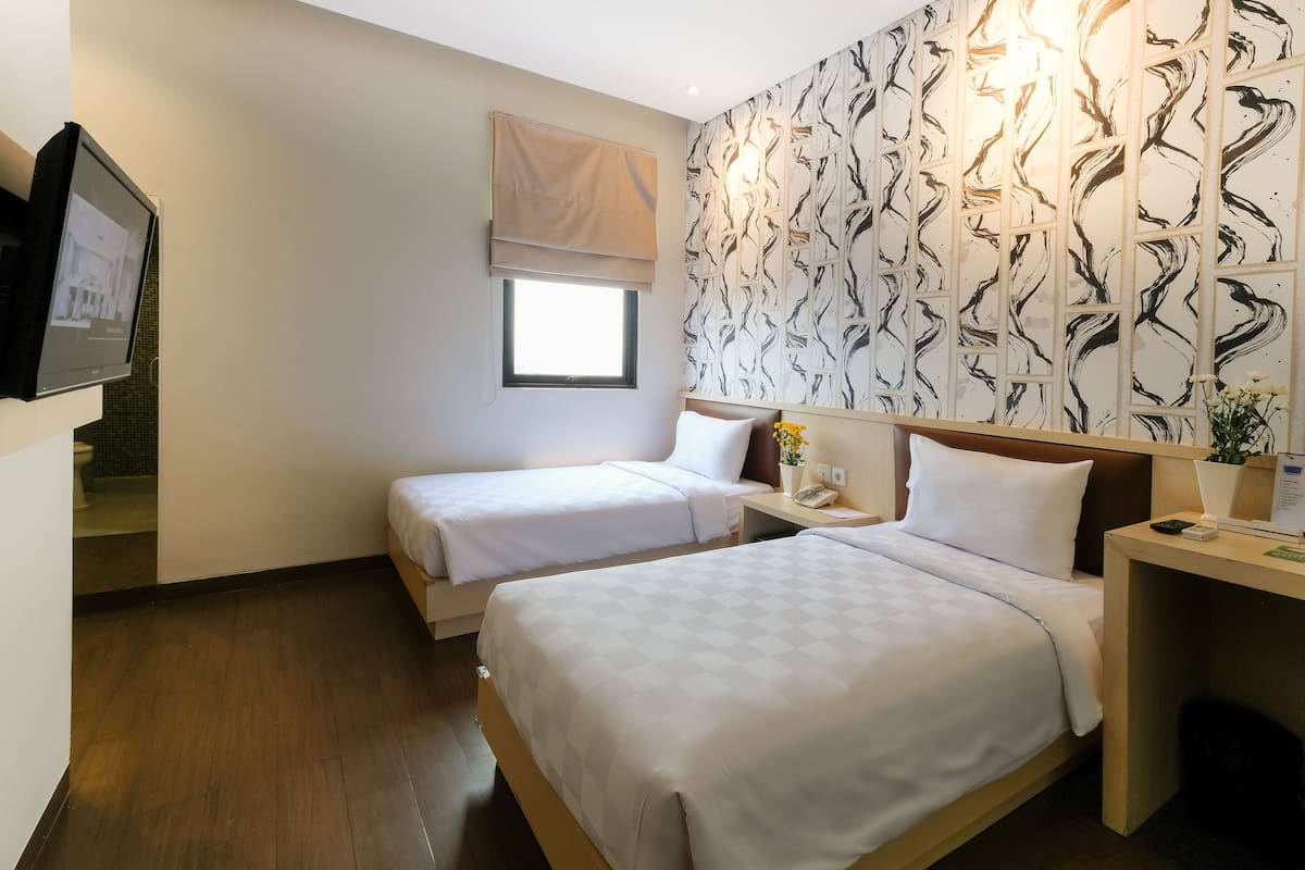 A hotel room with two beds, a small bedside table with a phone, a wall-mounted TV, and a window with a beige blind.