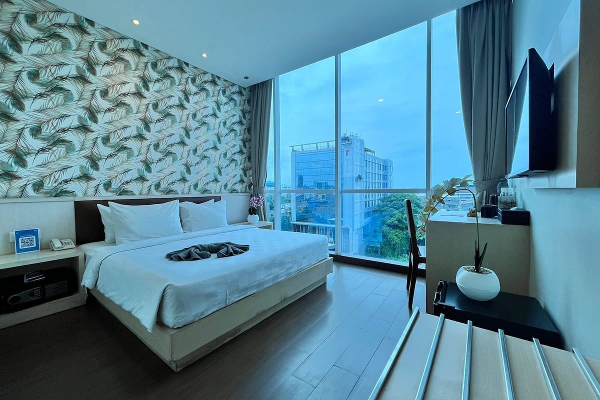 A modern hotel room with a large bed, a nightstand with a phone, a desk with a lamp, and a large window offering a city view.