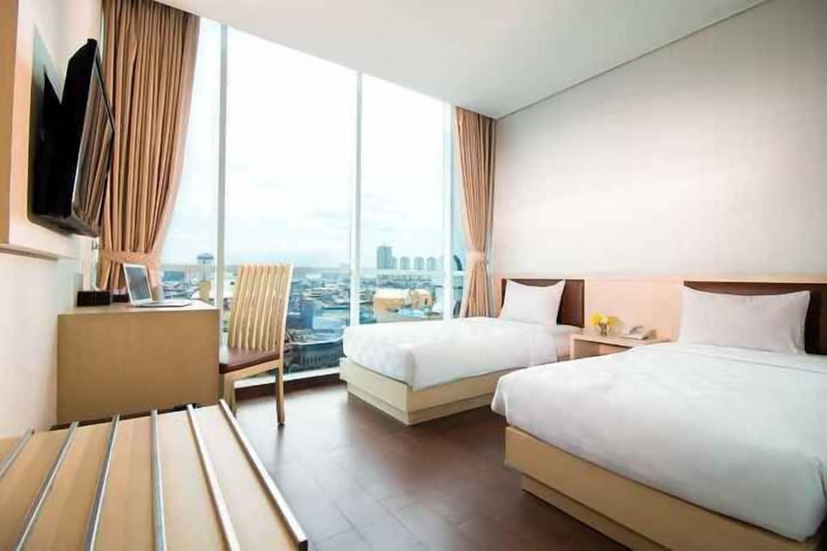 A hotel room with two beds, a desk, a chair, and a large window with a city view.