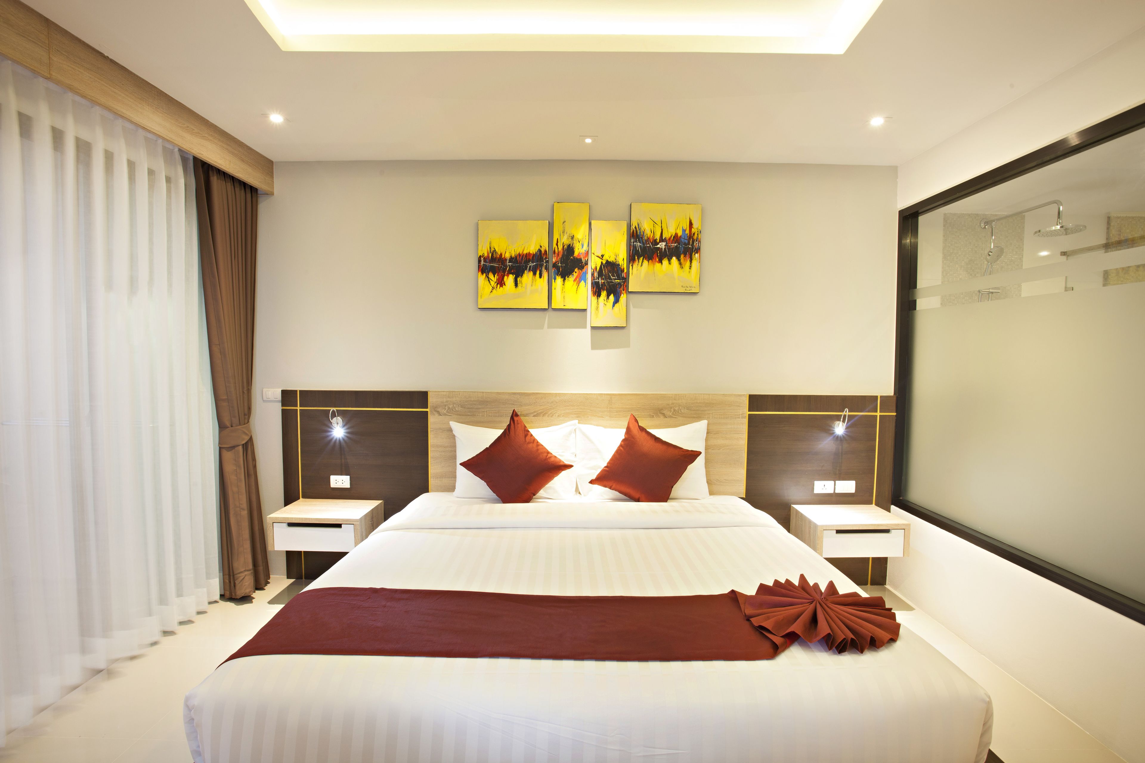 A modern hotel room with a large bed, two bedside tables, a nightstand, a large mirror, and three paintings on the wall.
