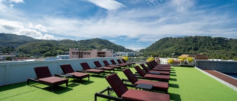 A rooftop lounge with rows of sun loungers on artificial grass, overlooking a mountainous landscape.