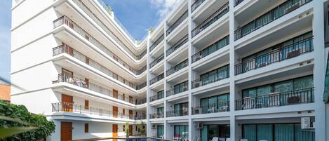 A multi-story hotel with a central swimming pool, balconies on each floor, and a clear sky.