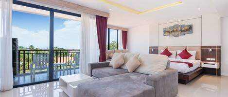 Une chambre d’hôtel moderne avec un canapé gris, un lit avec des oreillers rouges et blancs, une table de chevet et une grande fenêtre avec des rideaux.
