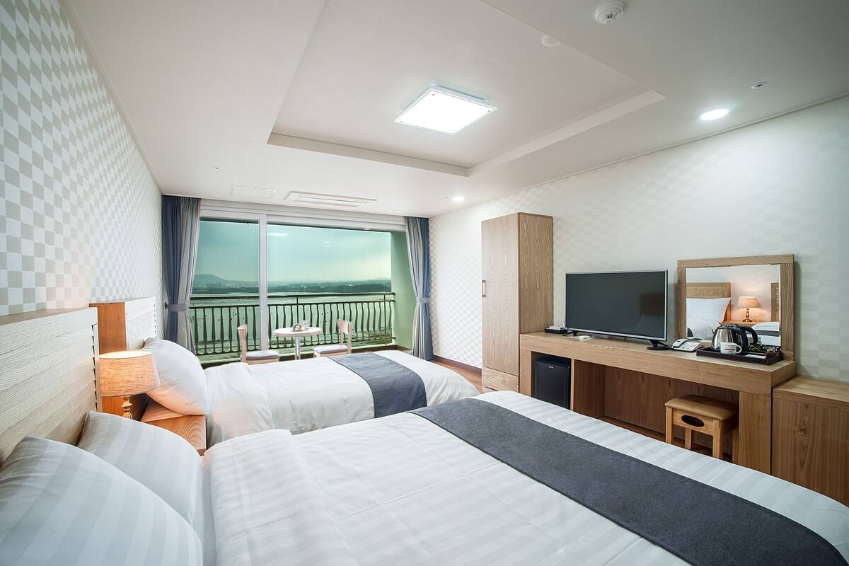 Ein Hotelzimmer mit zwei Betten, einem Schreibtisch mit Fernseher, einem kleinen Hocker und einem Balkon mit Blick.