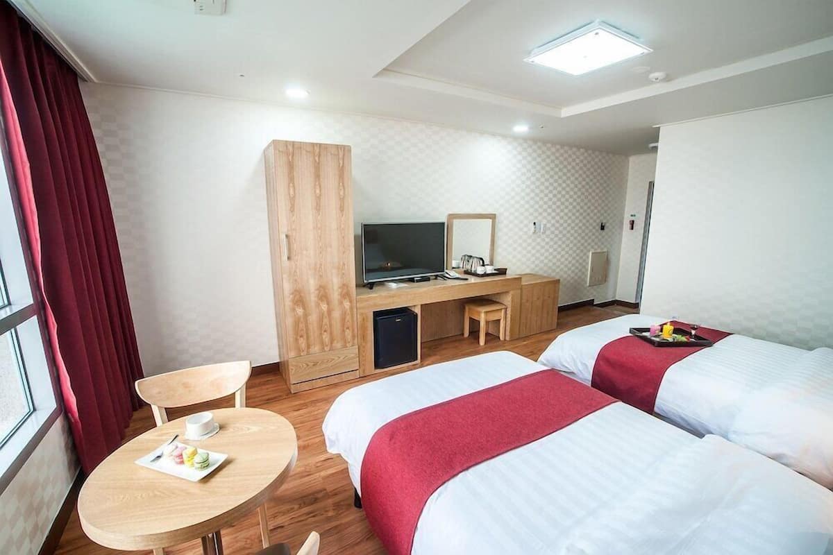 Ein Hotelzimmer mit zwei Betten, einem Schreibtisch mit Fernseher, einem kleinen Tisch mit Teller und einem Fenster mit Vorhängen.