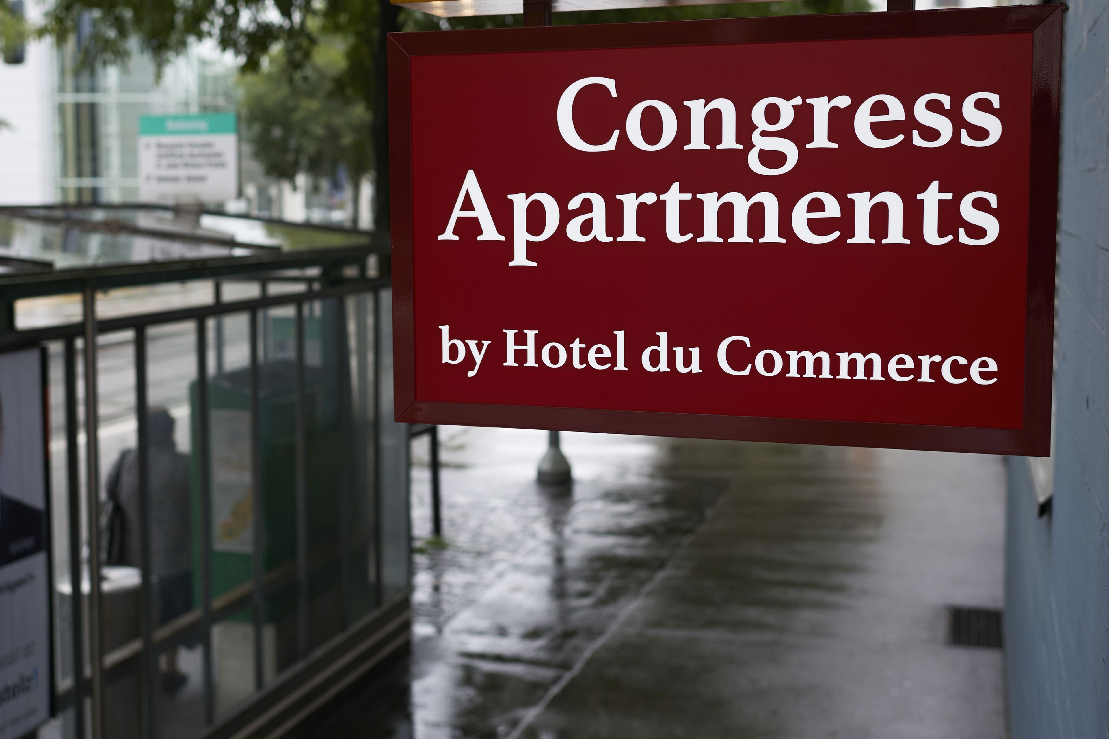 A sign for "Congress Apartments by Hotel du Commerce" with a metal railing and a wet pavement in the background.