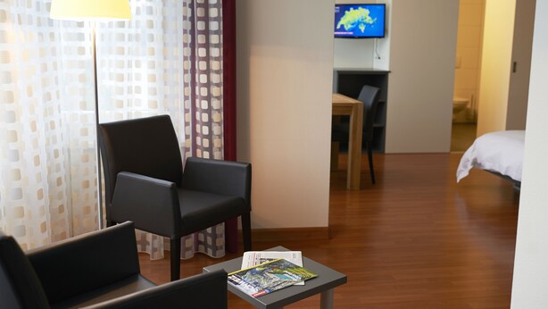 A hotel room with a bed, a desk, a chair, a lamp, and a TV. - Congress Apartments by Hotel du Commerce (Basel)