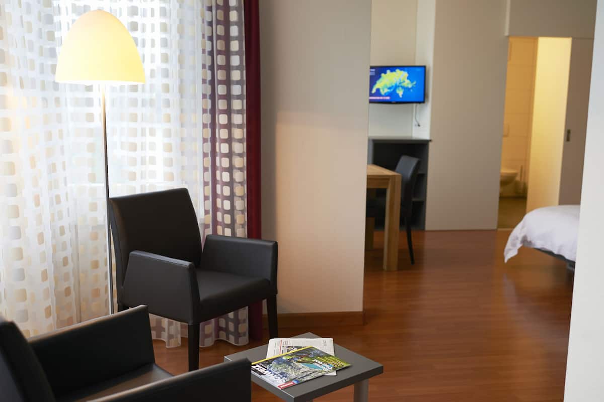 Quarto de hotel com cama, escrivaninha, cadeira, abajur e televisão.