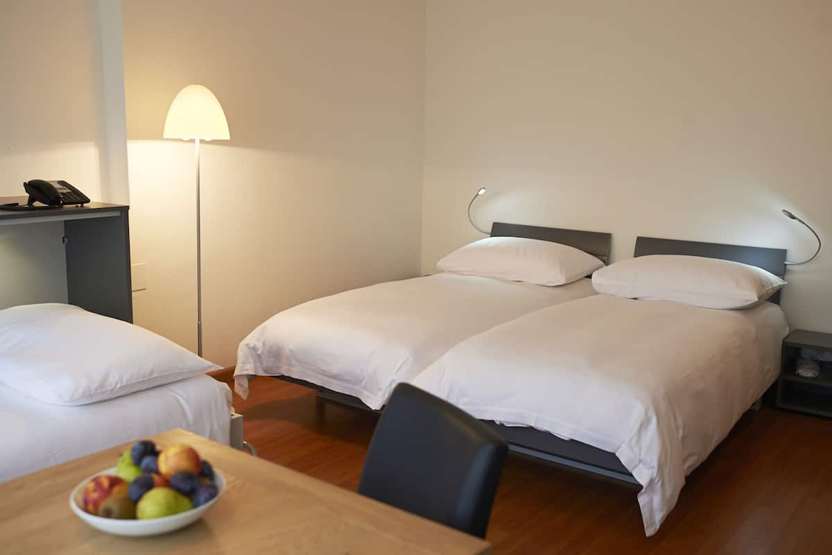 Quarto de hotel com duas camas, uma mesa de cabeceira com abajur, uma pequena escrivaninha com telefone e uma fruteira sobre a mesa.