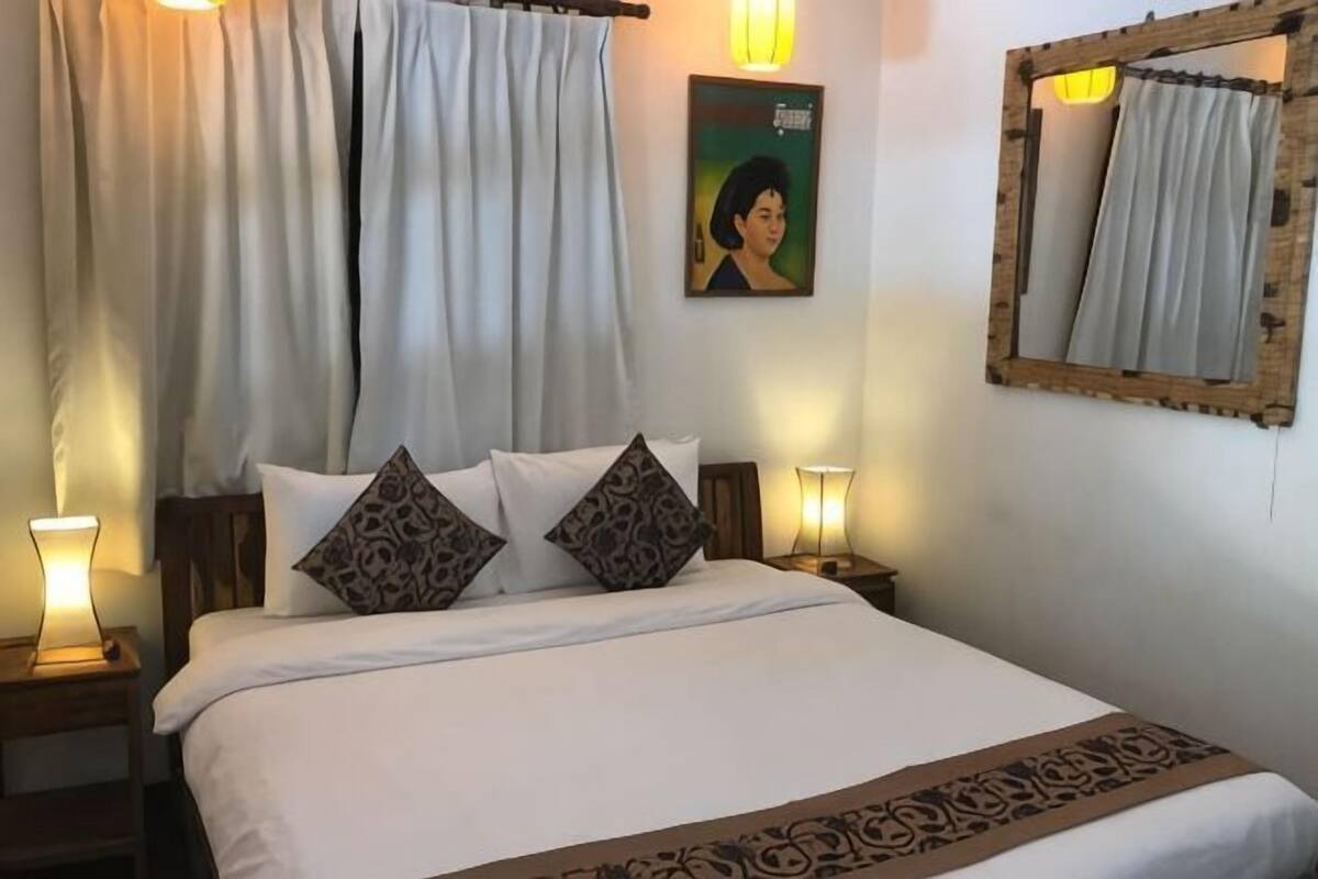 Una cama bien tendida con ropa blanca y almohadas estampadas, dos lámparas de noche, un cuadro en la pared y un espejo con marco de madera.