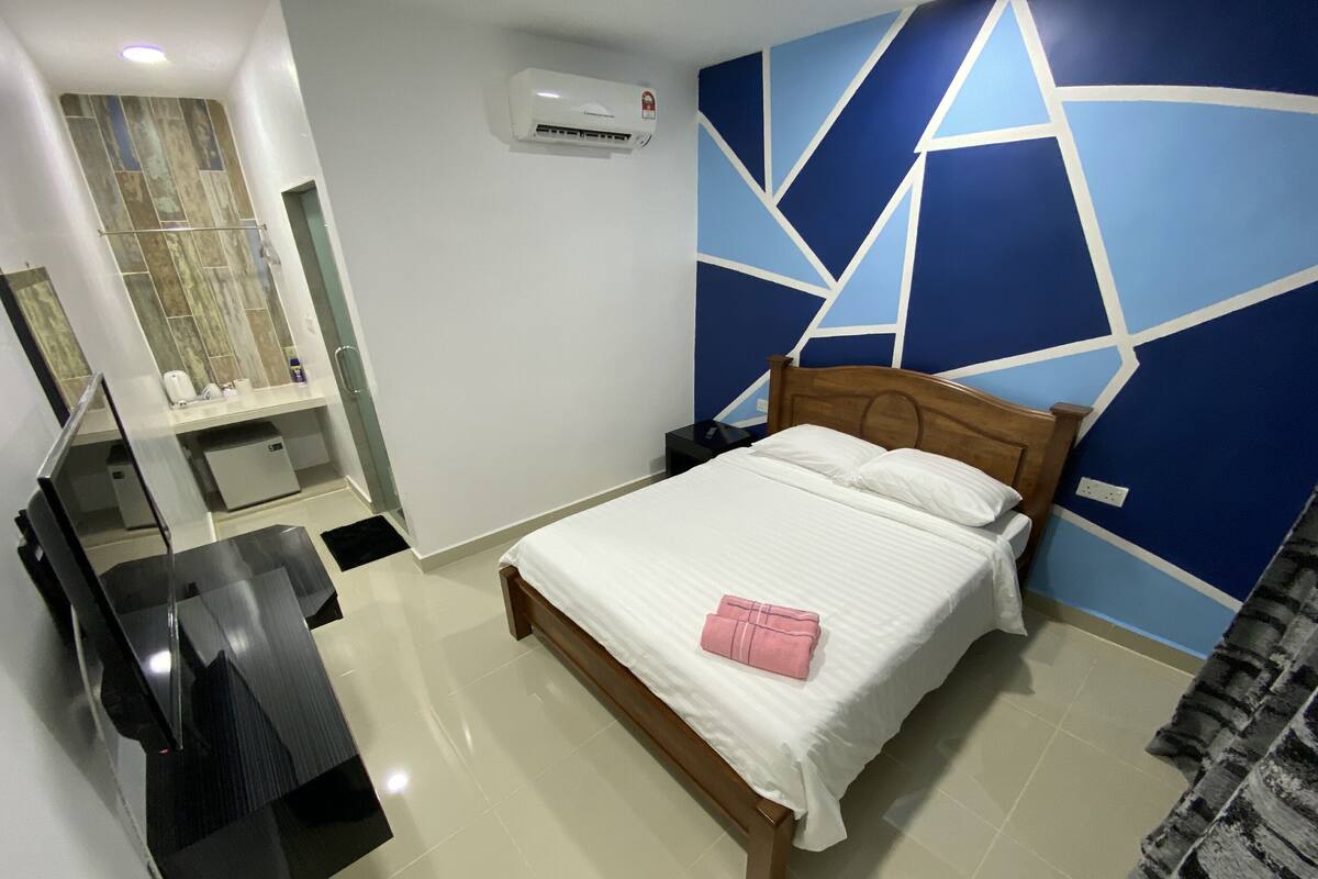 A hotel room with a bed, a desk, a mirror, and a wall-mounted air conditioner.