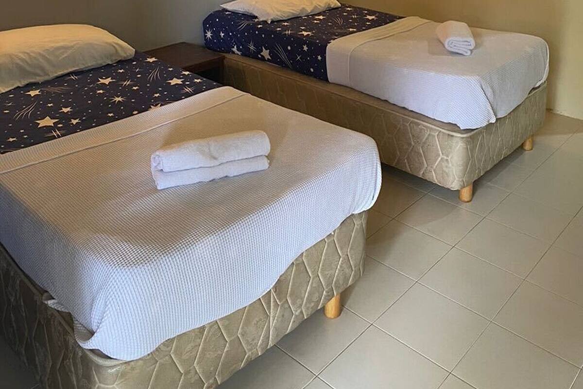 A hotel room with two beds, each with a white blanket and a star-patterned bedspread. There is a wall-mounted air conditioner and a wall sconce.