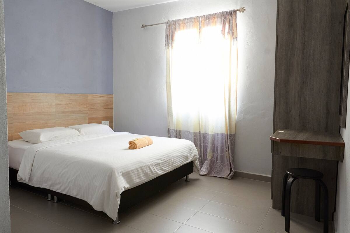 A bedroom with a bed, a wooden headboard, a window with curtains, a small stool, and a desk area.
