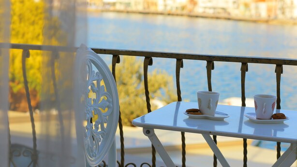 A balcony with a table set for two, overlooking a river and buildings. - Huri & Nuri Hotel (Foça)