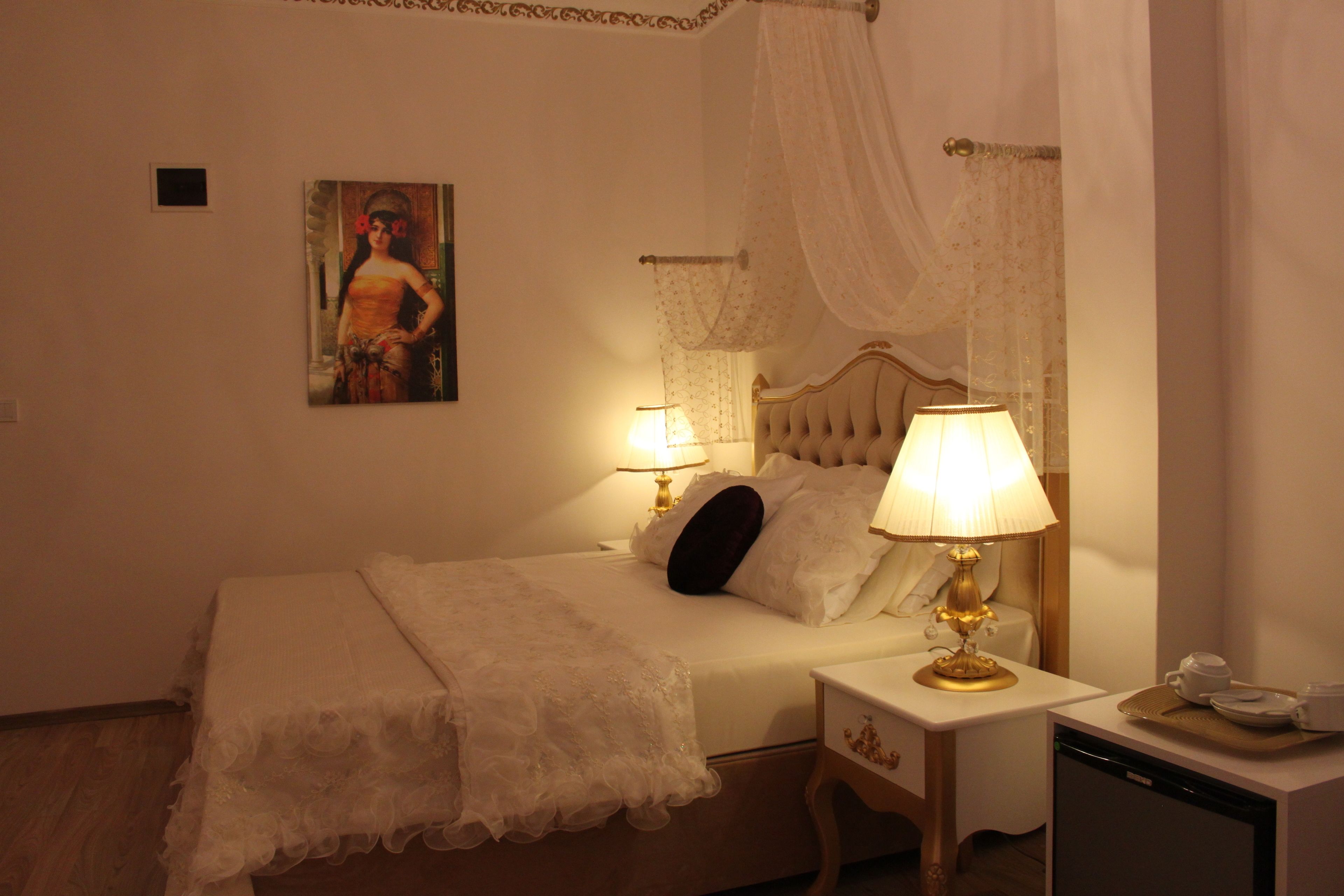 A neatly made bed with a white bedspread and pillows, a bedside table with a lamp, a small bedside table with a cup and saucer, and a painting on the wall.
