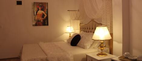A neatly made bed with a white bedspread and pillows, a bedside table with a lamp, a small bedside table with a cup and saucer, and a painting on the wall.