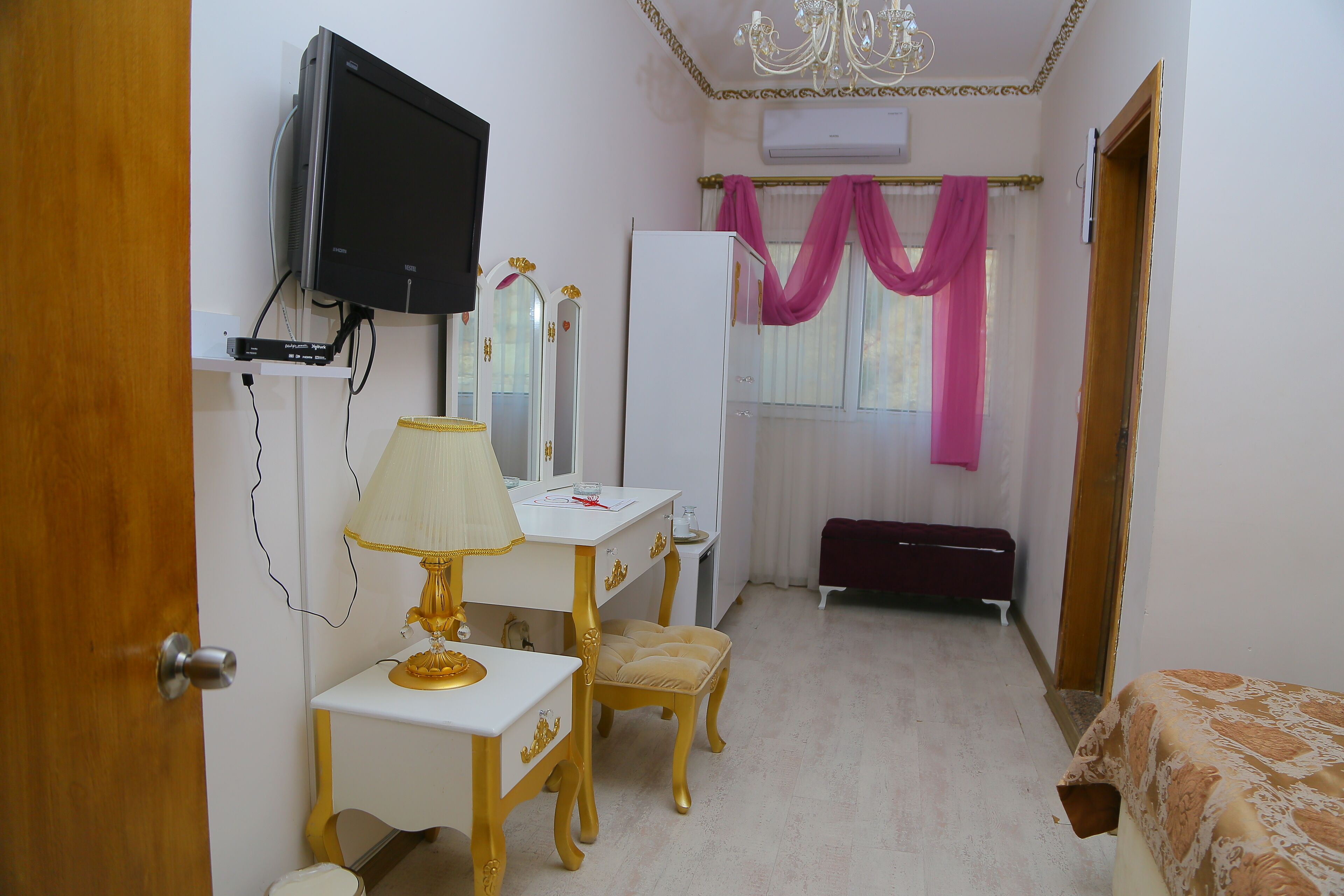 A bedroom with a TV mounted on the wall, a vanity with a mirror, a bedside table with a lamp, a bed with a patterned bedspread, and a window with pink curtains.