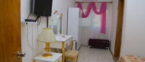 A bedroom with a TV mounted on the wall, a vanity with a mirror, a bedside table with a lamp, a bed with a patterned bedspread, and a window with pink curtains.