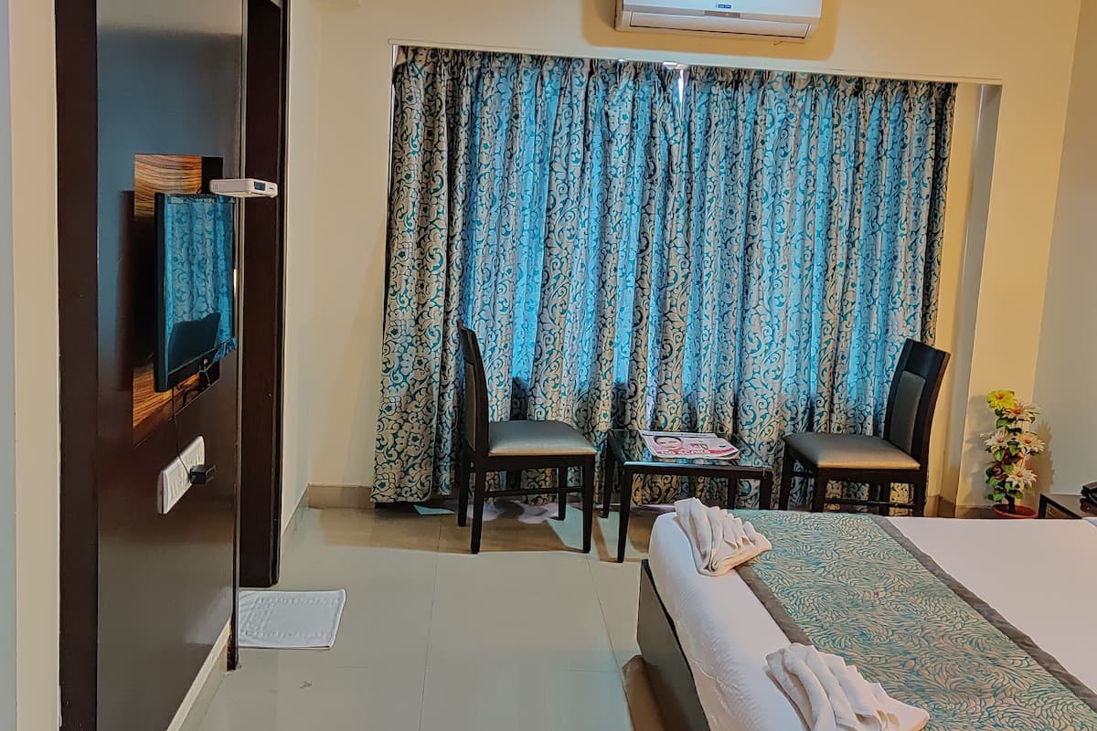 A hotel room with a bed, two chairs, a small table, a TV, and an air conditioning unit.