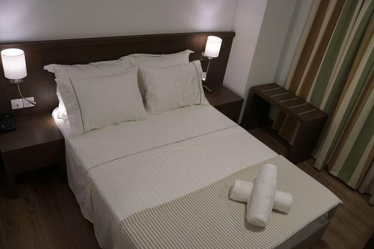 A hotel room with a bed, two bedside lamps, a nightstand, a small table, and a curtain.