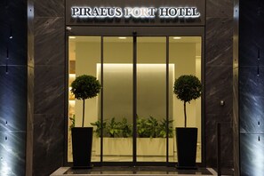 El exterior del Hotel Piraeus Port, con una fachada moderna, puertas de cristal grandes y dos macetas con plantas en la entrada.