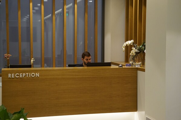 Un mostrador de recepción con la etiqueta "RECEPTION", una persona sentada detrás, un jarrón con flores blancas y paneles de madera verticales al fondo.