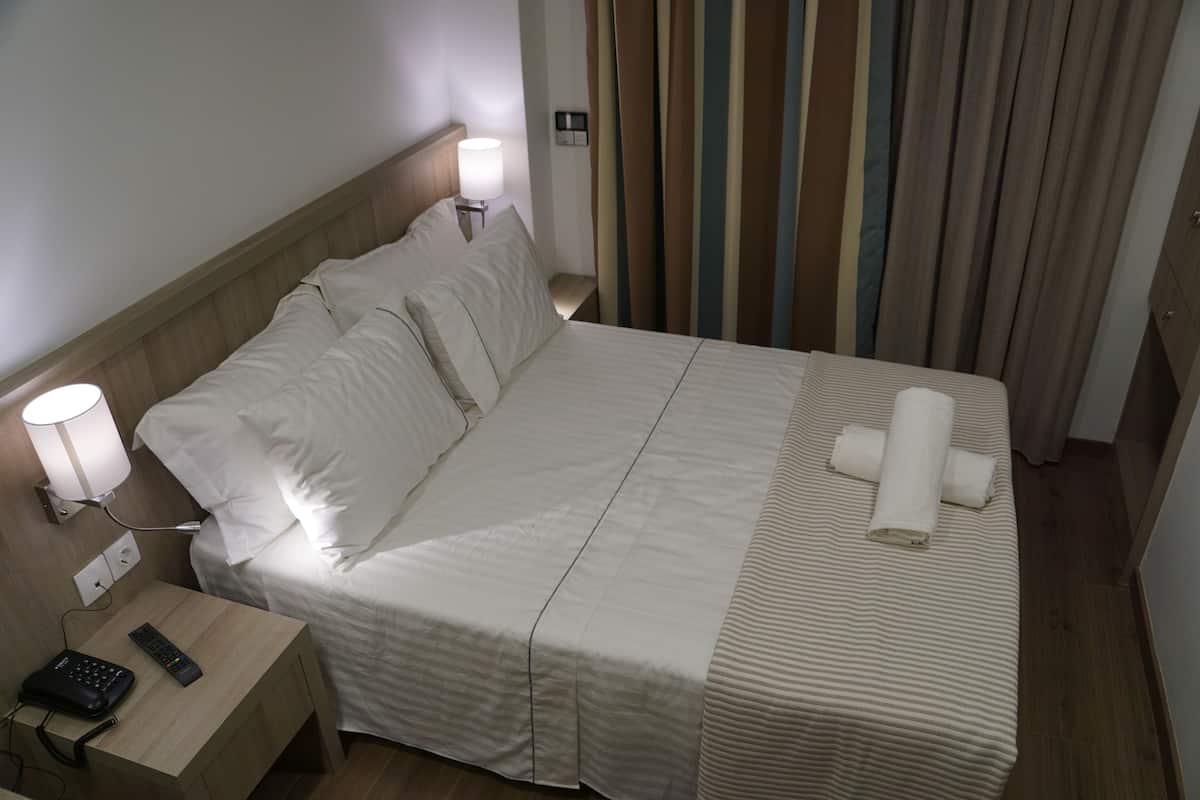 A hotel room with a neatly made bed, a bedside table with a lamp, and a remote control.