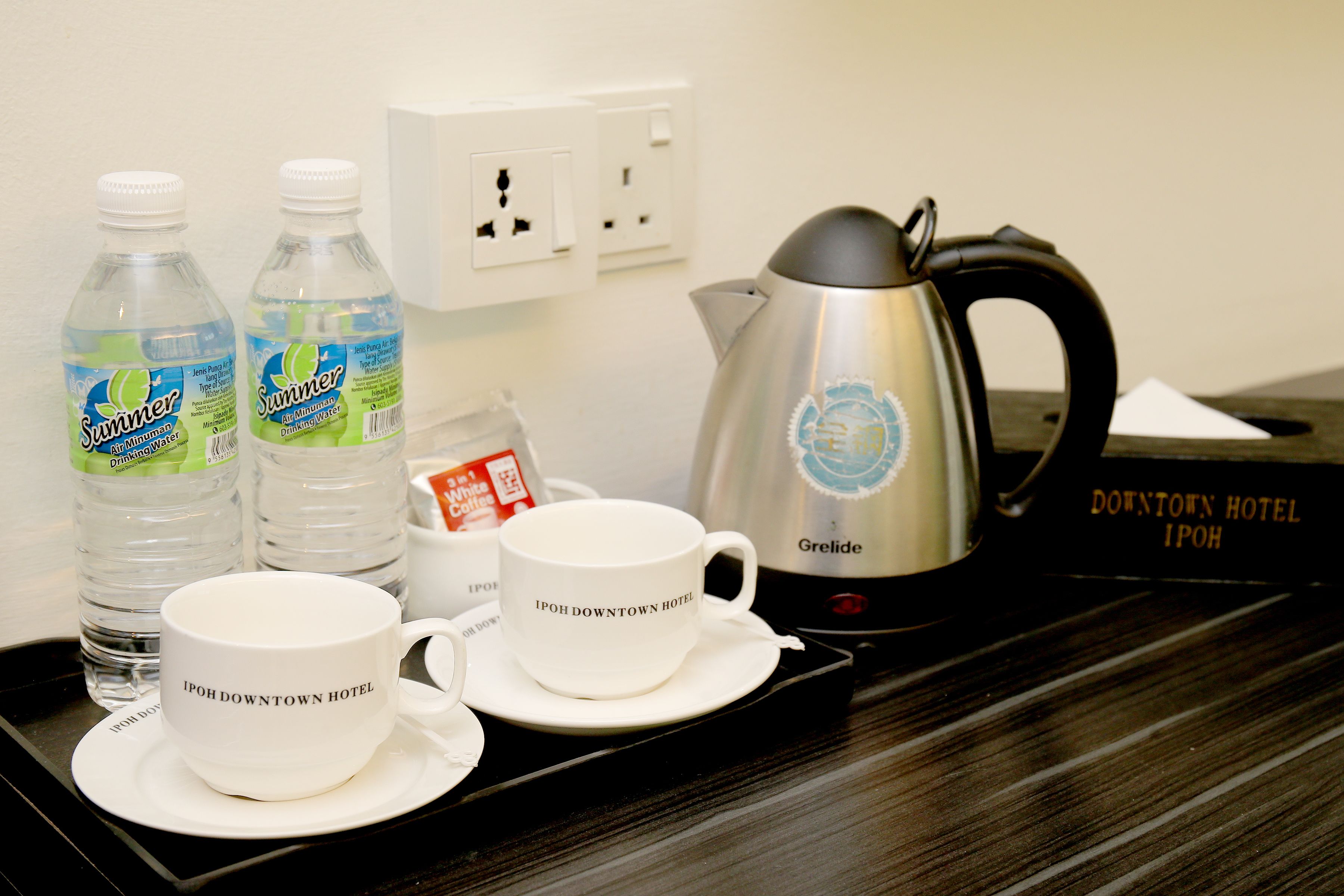 A tray with two white cups labeled "Ipoh Downtown Hotel," a silver electric kettle, and two bottles of water.