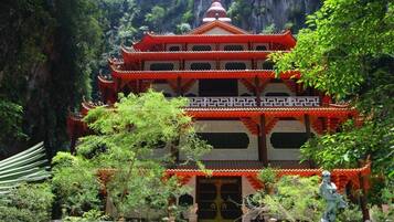 A traditional multi-tiered temple with a red roof, surrounded by lush greenery and a rocky cliff backdrop.
