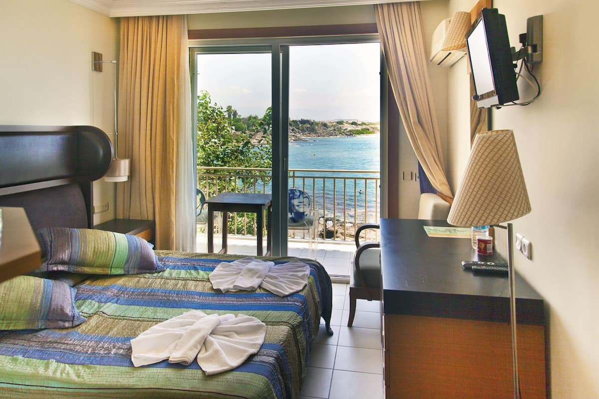 A hotel room with a bed, a desk, a chair, a TV, and a view of the sea.