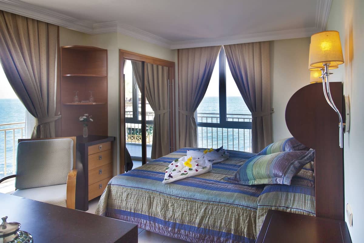 A hotel room with a bed, a desk, a chair, a lamp, and a view of the sea.