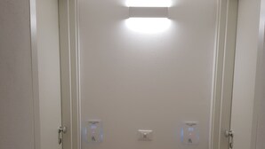 A narrow hallway with white walls and wooden flooring. There are three doors on the left, each with a doorbell and a light switch. The hallway has a ceiling light.