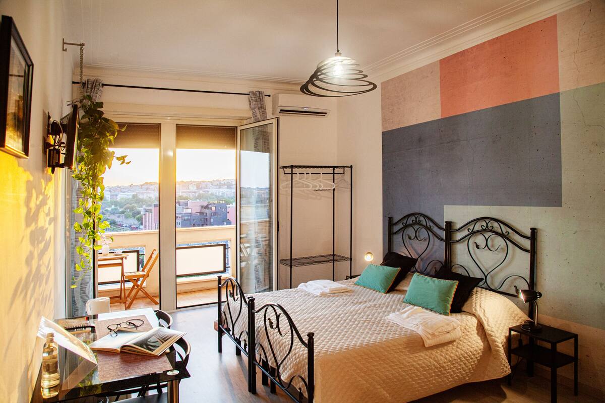 A bedroom with a bed, bedside table, lamp, and a view through a sliding door to an outdoor balcony.