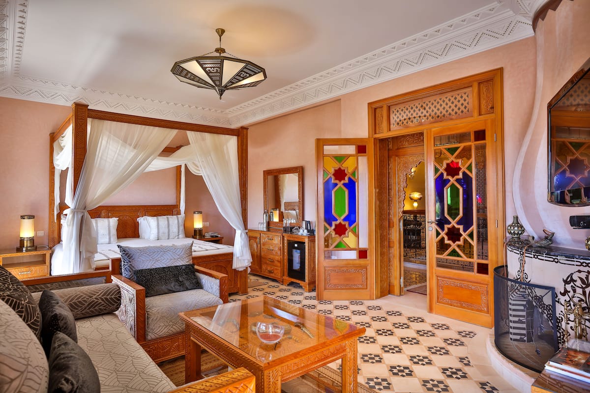 A traditional room with a four-poster bed, a sitting area with a glass-top table, and a decorative door with stained glass.